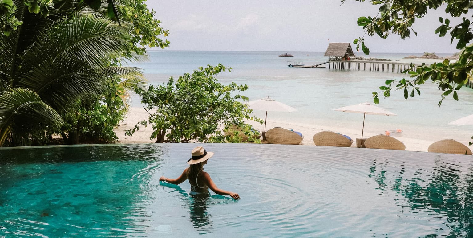 A woman in a sun hat wading in an infinity pool that overlooks a tropical beach